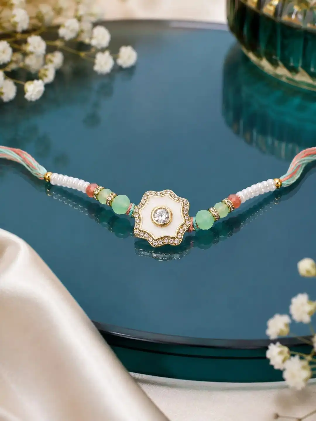 RHOSYN floral rakhi features a white and gold centerpiece with pastel green beads on an adjustable thread.