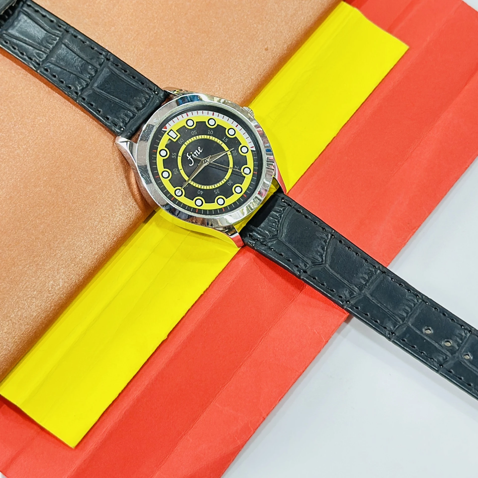 Fine" brand wristwatch featuring a black strap and a dial with yellow and black accents.