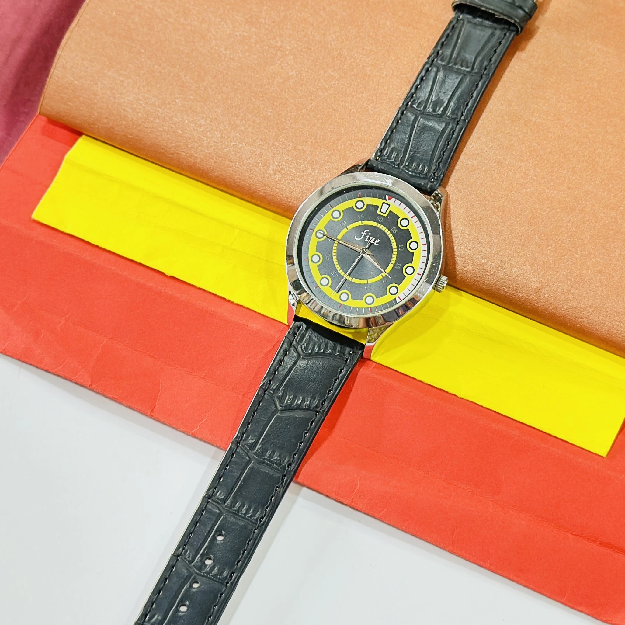Fine" brand wristwatch featuring a black strap and a dial with yellow and black accents.