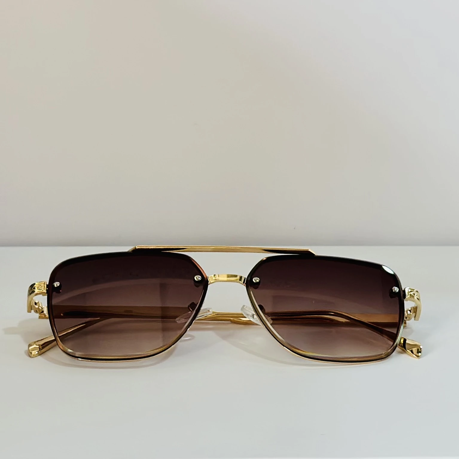 pair of rimless gradient brown aviator sunglasses with a gold metal frame and ornate side details.
